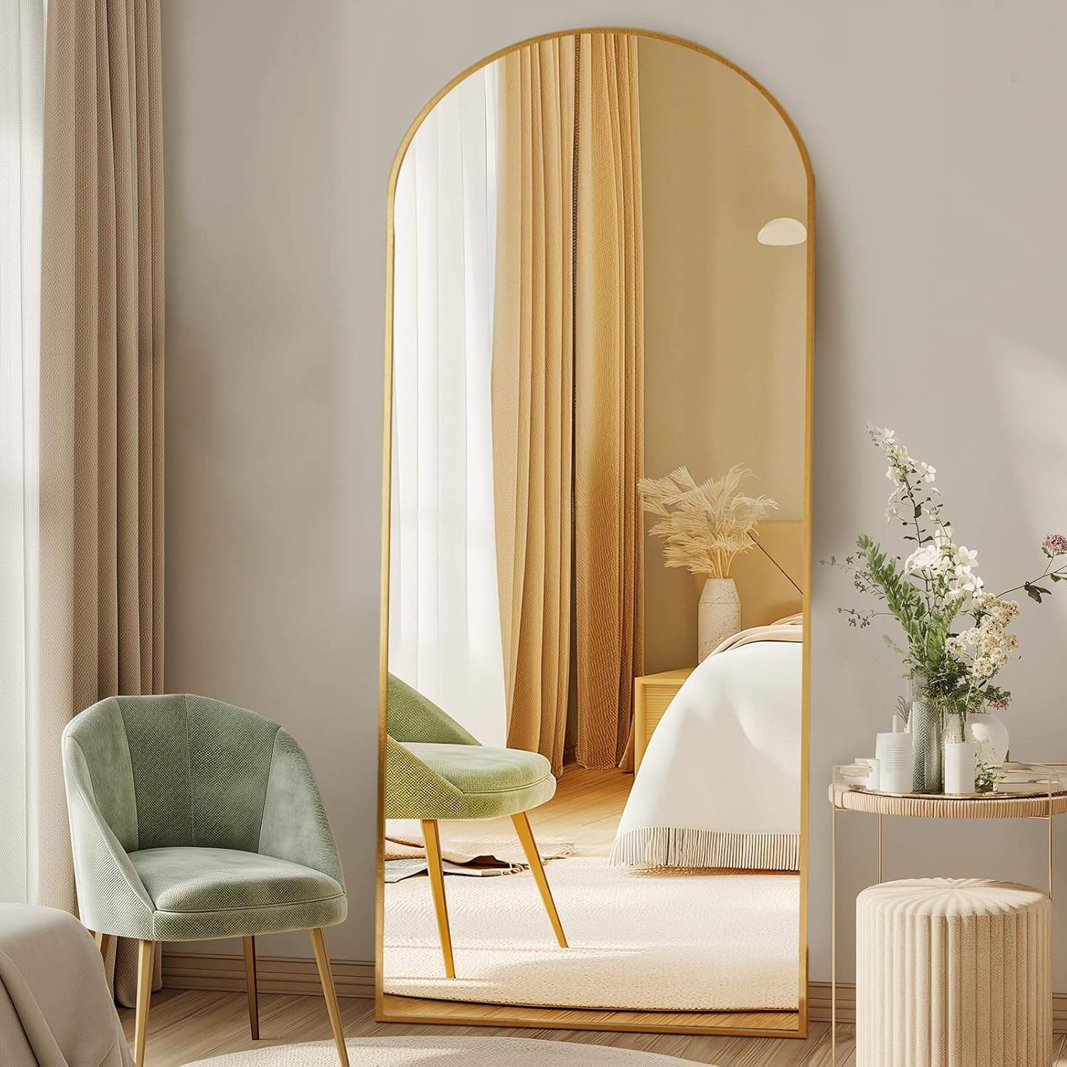 Gold arched full length mirror styled in a warm neutral bedroom with soft curtains, sage chair, flowers, wood flooring, and bright natural light.