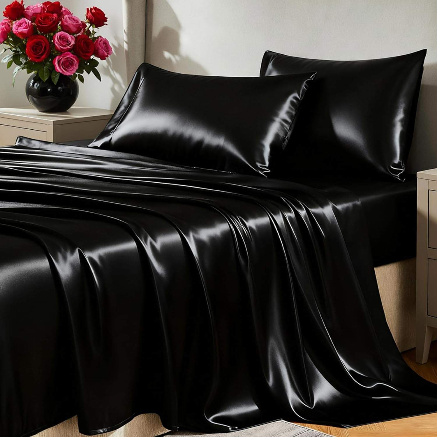 Black satin sheets and pillows styled on a modern bed with cream nightstands, bright window light, glossy fabric texture, and red roses for a cozy dark bedroom mood.