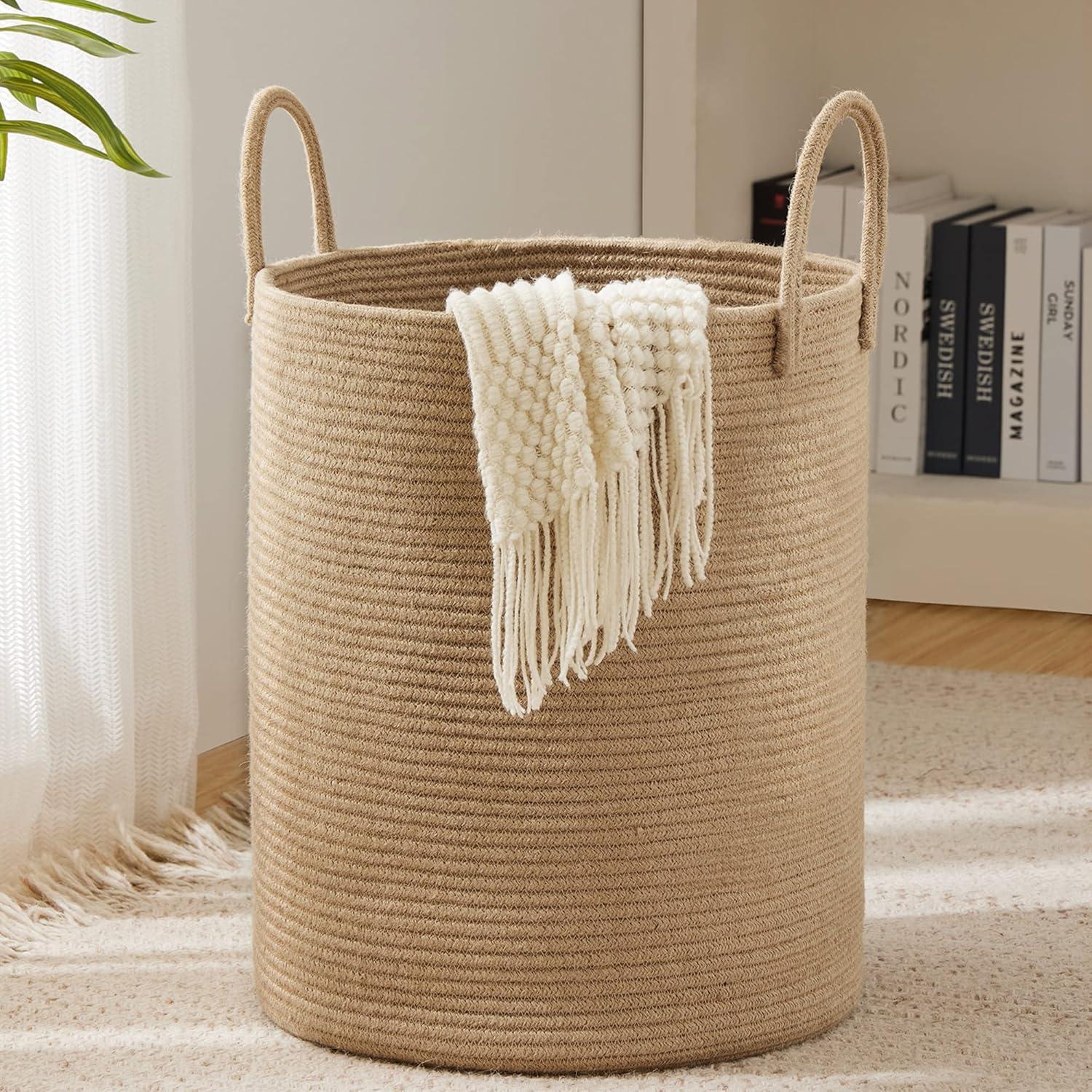 Tall jute rope laundry hamper with loop handles holding a cream throw in a bright neutral bedroom with soft curtains, books, plant leaves, and warm natural textures.