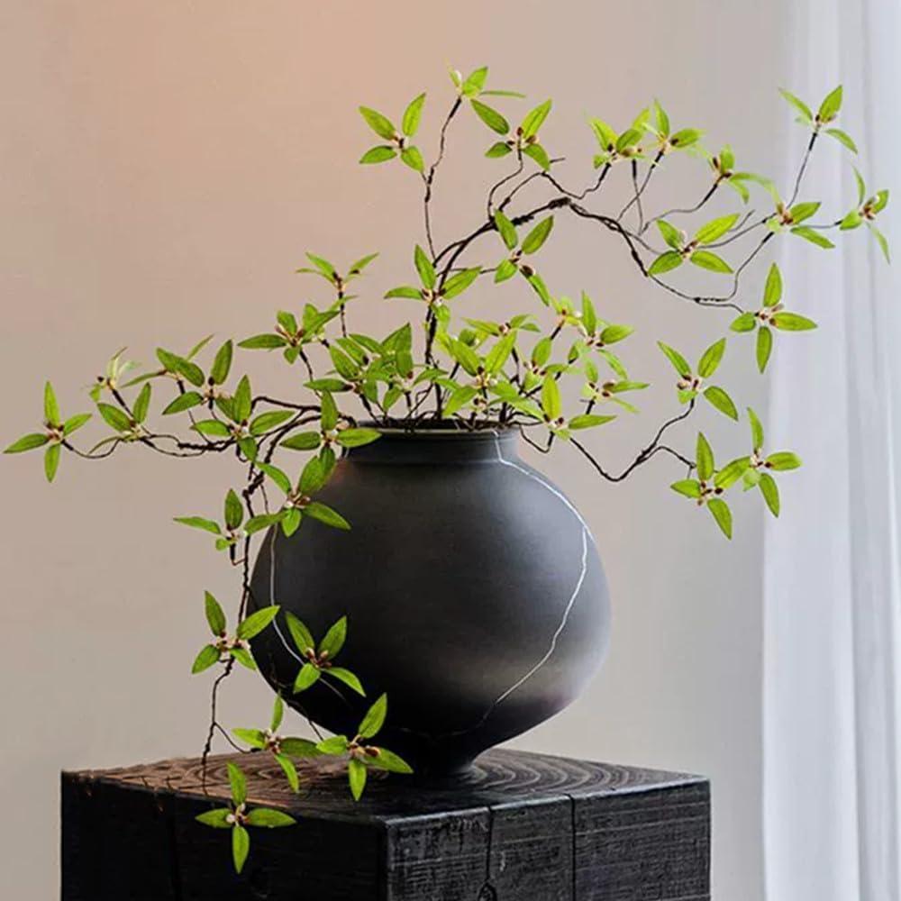Faux greenery stems arranged in a dark ceramic vase on a wood console beside soft window light, styled for calm neutral home decor.