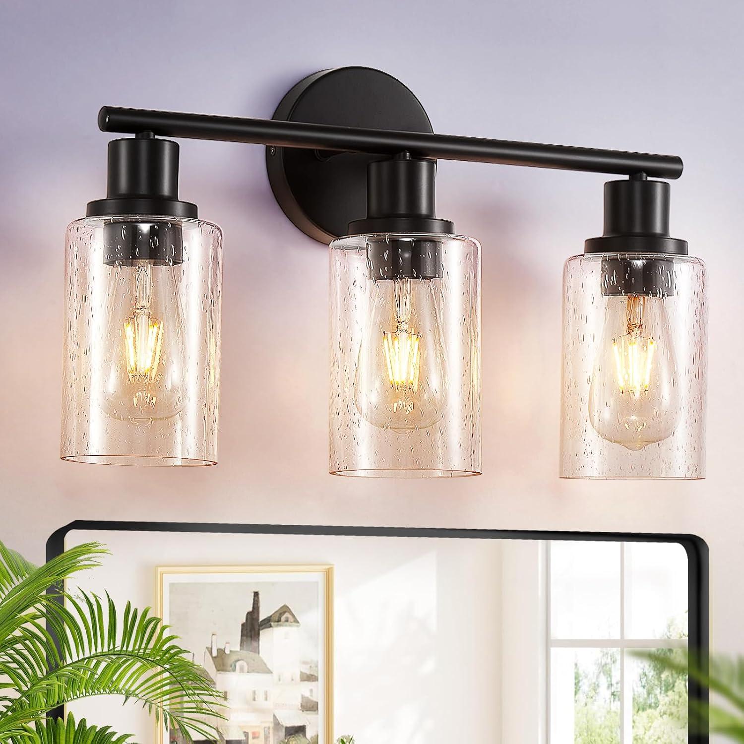 Matte black three-light vanity fixture with clear glass shades glowing above a bathroom mirror, styled with bright natural light, soft neutral walls, framed art, and green plant leaves.