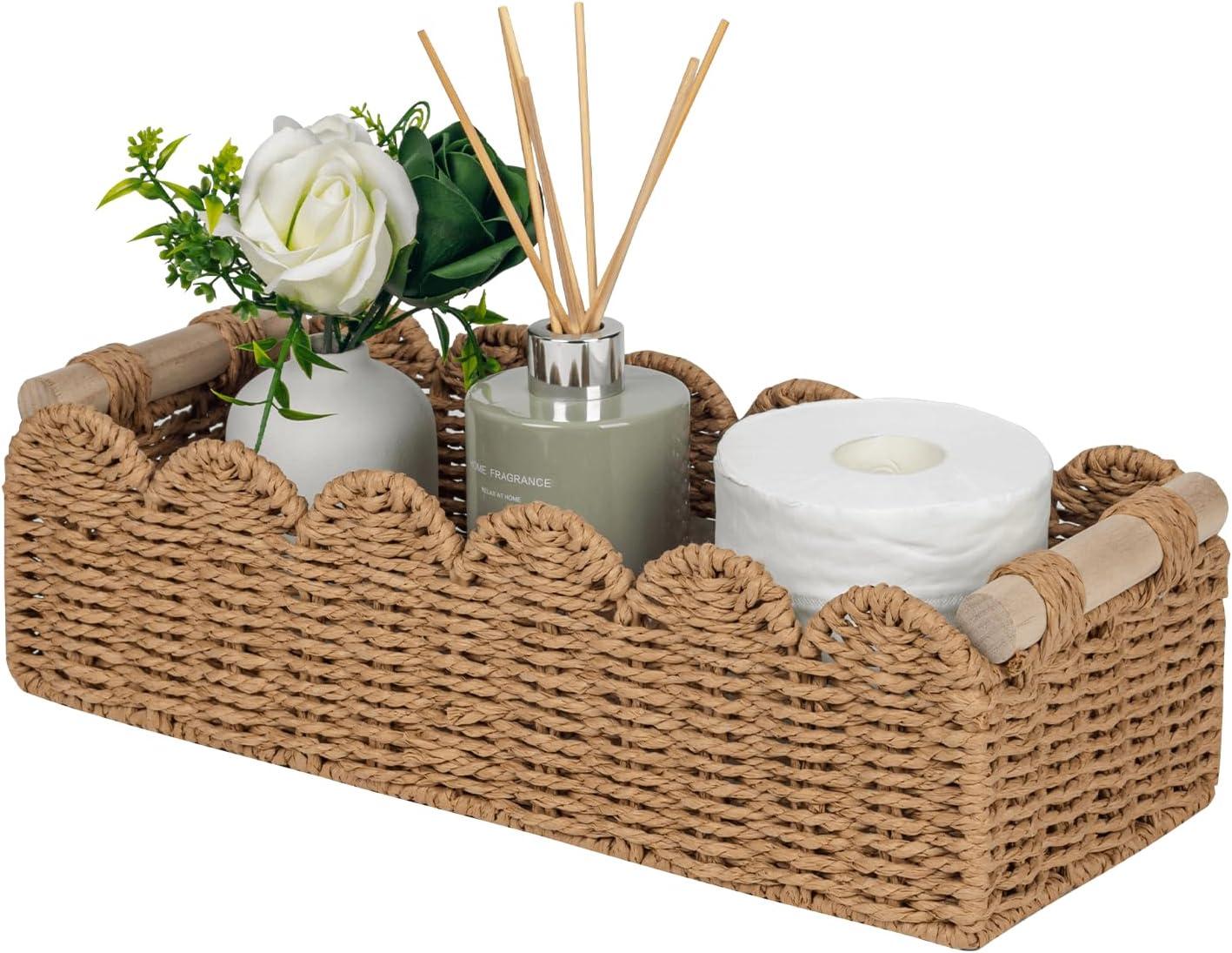 Neutral woven scalloped basket with wooden handles styled with toilet paper, reed diffuser, ceramic vase, white rose, and greenery for calm bathroom storage decor.
