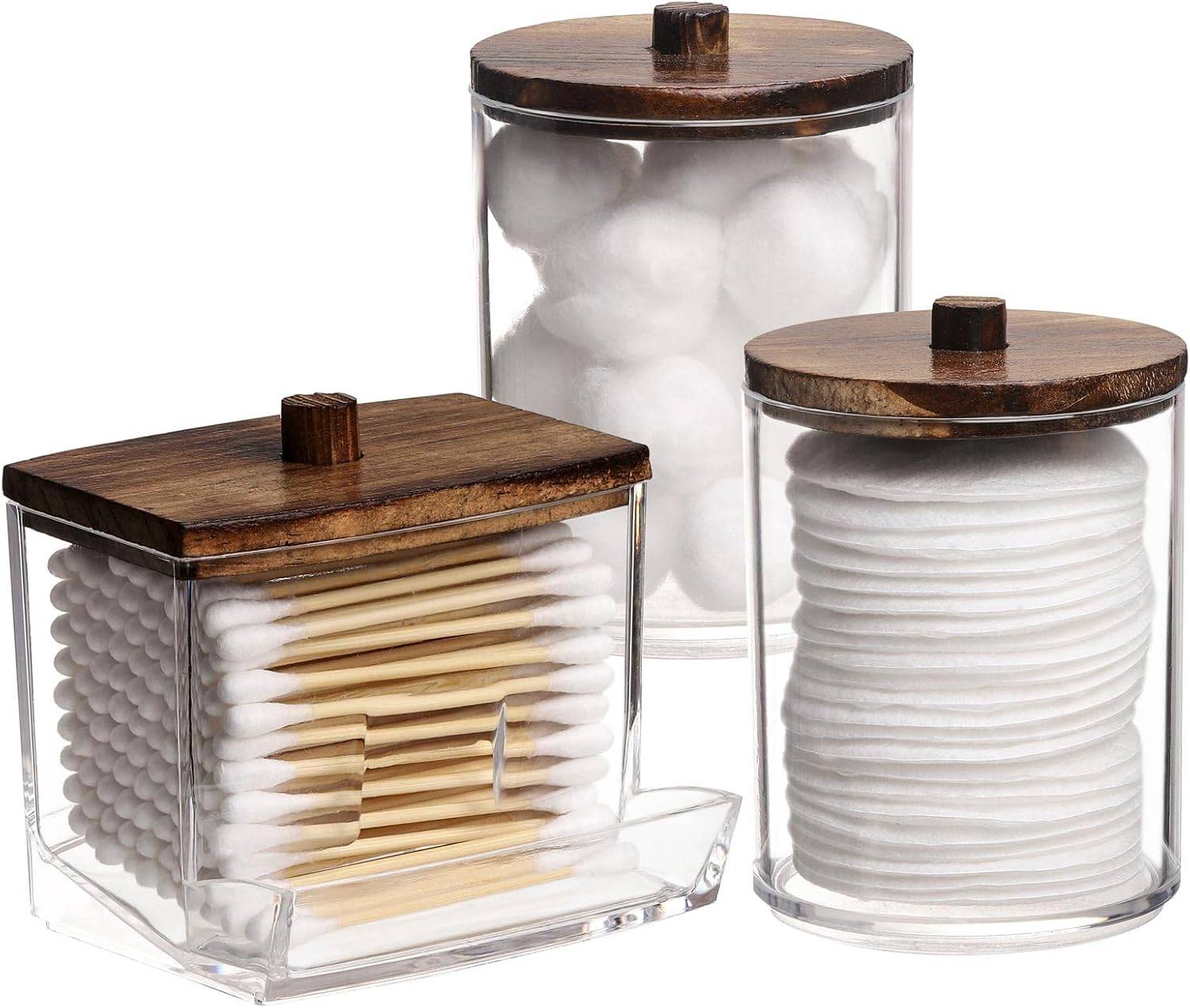 Clear bathroom apothecary jars with warm wood lids holding cotton balls, swabs, and pads on a bright neutral vanity with cozy minimalist style.