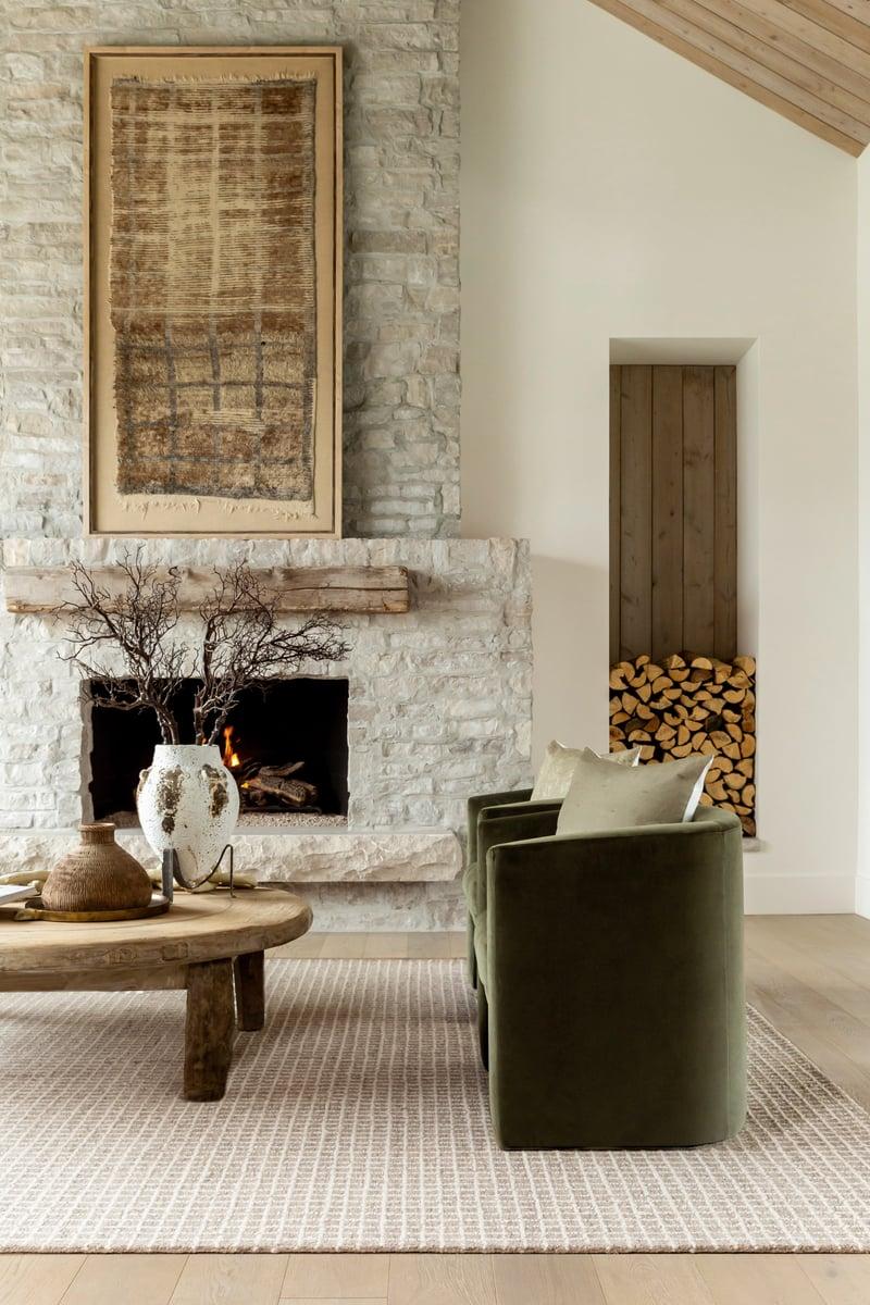 Cozy neutral living room with a stone fireplace, textured area rug, olive lounge chair, wood coffee table, ceramic vase, and stacked firewood in soft natural light.