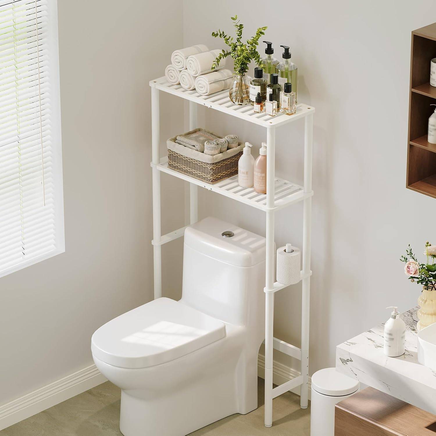 Bright neutral bathroom with a white over toilet storage shelf styled with rolled towels, a woven basket, bottles, greenery, and soft natural light.