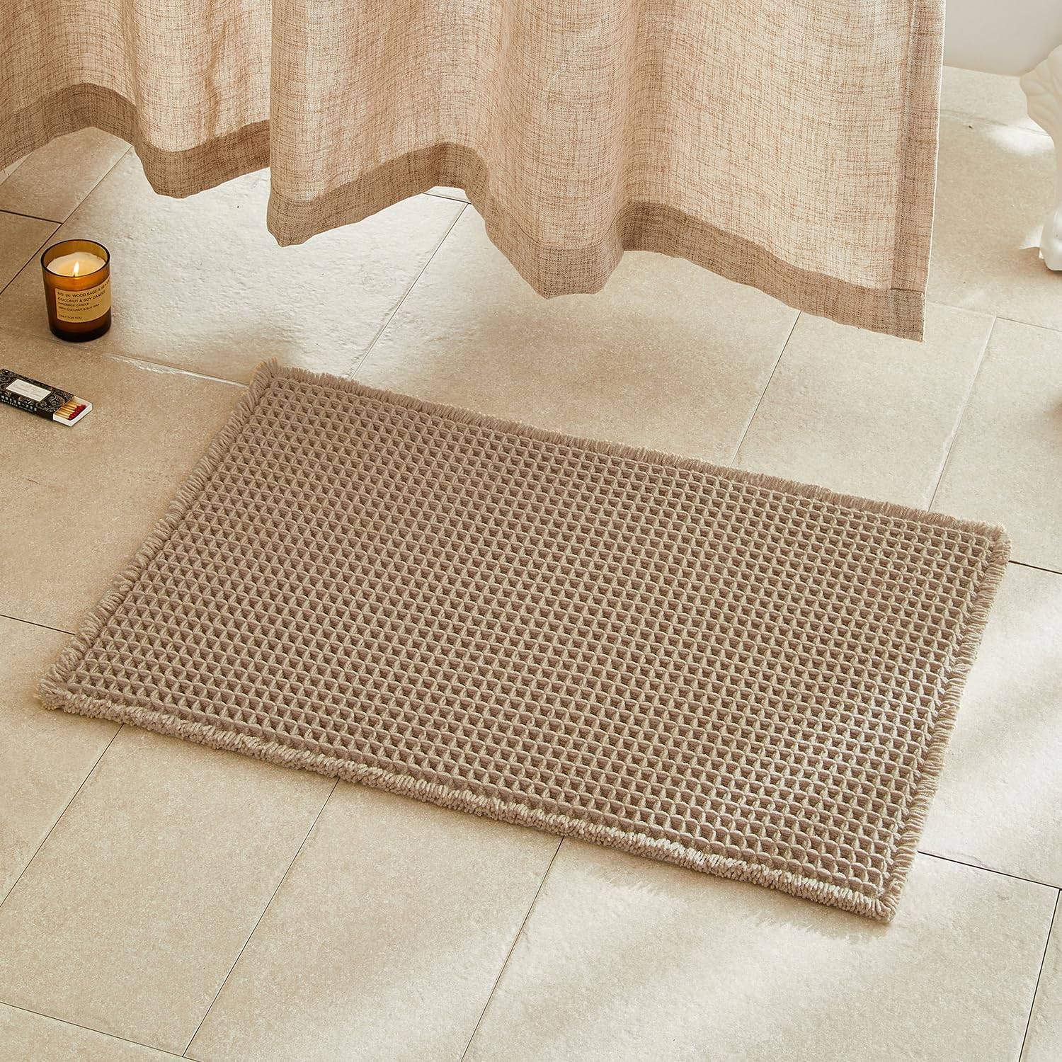 Khaki waffle bath mat on beige tile beside a soft linen shower curtain, amber candle, and warm natural light for cozy neutral bathroom decor.