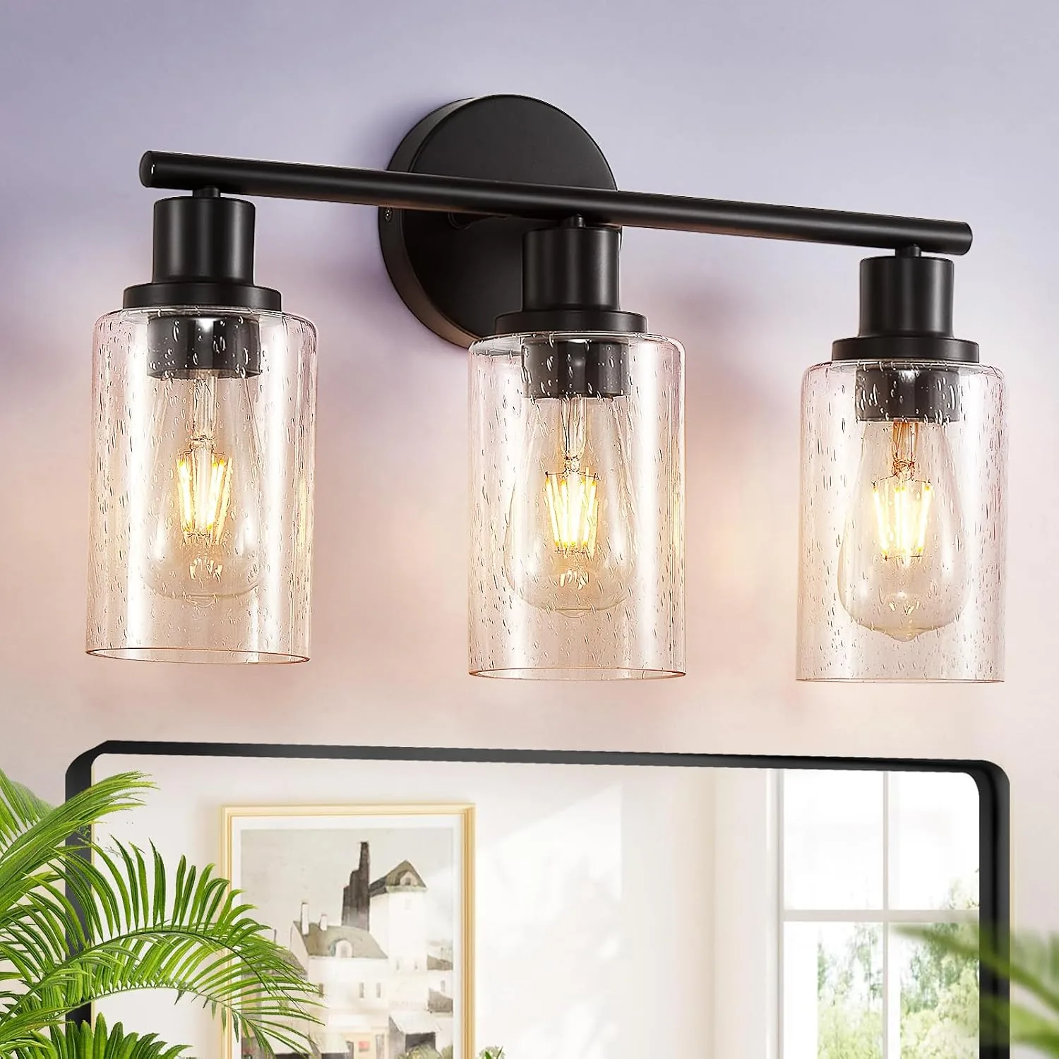 Matte black three-light vanity fixture with clear glass shades glowing above a bathroom mirror, styled with bright natural light, soft neutral walls, framed art, and green plant leaves.
